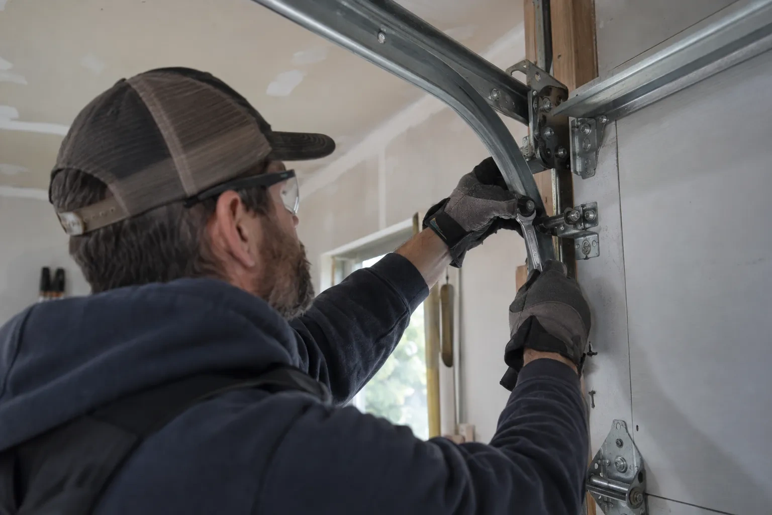 Technician in a mesh cap and safety glasses holding a wrench to a bracket at the bend of the metal door track. — Buchanan