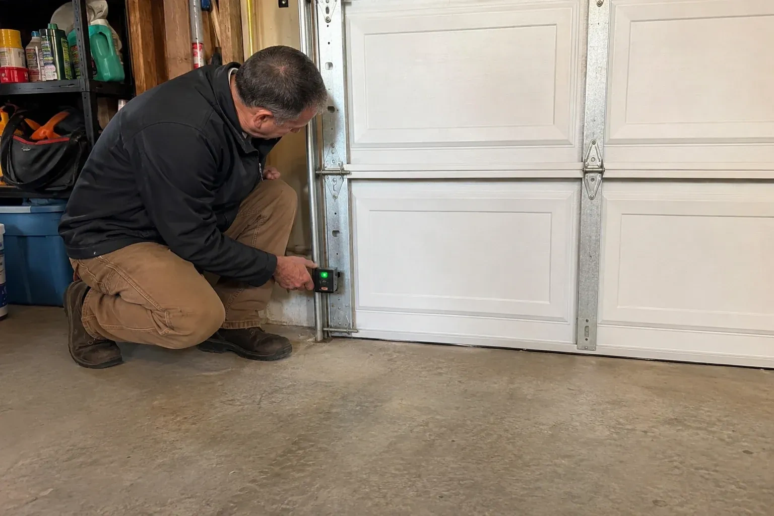 Technician kneeling beside a white paneled garage door, holding a small black box with a green light against the vertical track. — Photo-Eye Sensor Alignment or Replacement, Hamilton